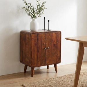 May include: A dark brown wooden cabinet with rounded corners and two doors featuring small black knobs. The cabinet stands on four tapered legs. On top sits a white ceramic vase containing green leafy branches and two thin black candle holders.