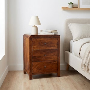 May include: A three-drawer wooden nightstand with a warm brown finish. The nightstand has a rounded top and small, dark knobs on each drawer. A small white lamp and two books sit on top. The nightstand is next to a bed with a neutral-colored headboard.