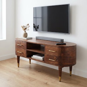 May include: A dark brown, oval TV stand with four drawers and gold handles. The stand has two open shelves and gold-tipped legs. A flat-screen TV is mounted above the stand, with a soundbar and remote control on top. A vase with dried flowers sits on the stand.