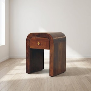May include: A dark brown wooden bedside table with a single drawer and a brass knob. The table has a curved top and a rectangular base. The dimensions are approximately 61 cm high, 51 cm wide, and 41 cm deep.