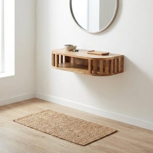 Op de afbeelding: Een aan de muur gemonteerde zwevende plank van lichtgekleurd hout, voorzien van een lattenstructuur aan de voorzijde en afgeronde hoeken. Op de plank staan een kleine witte kom en een rechthoekig houten dienblad. Een ronde spiegel hangt erboven.