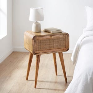 May include: A light brown wooden nightstand with four tapered legs and a single drawer featuring a woven cane front panel. A small beige table lamp with a white fabric shade and a closed book are placed on the flat top surface of the nightstand.