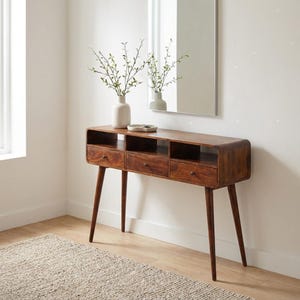 Könnte beinhalten: Eine Holzkonsole im modernen Stil der Mitte des Jahrhunderts mit drei Schubladen und offenen Regalen. Der Tisch hat konische Beine und eine geschwungene Front. Eine weiße Vase mit Grün und eine kleine Schale stehen oben. Ein Spiegel ist im Hintergrund.
