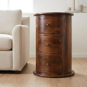 Op de afbeelding: Een donkerbruine houten nachtkast met drie lades en een afgeronde vorm. De nachtkast heeft een ronde basis en bovenkant, met kleine, ronde knoppen op elke lade. De houtnerf is zichtbaar en de afwerking is glanzend.