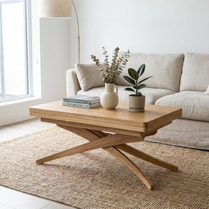 May include: A rectangular light oak coffee table featuring a unique scissor-style cross-leg base design. The table surface is smooth and flat, providing a stable platform for decorative items like books, a ceramic vase, and a small potted plant.