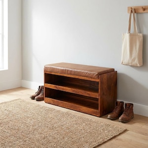 May include: A wooden shoe bench with a brown leather cushion top and two open shelves. Two pairs of brown leather boots are placed near the bench. A beige tote bag hangs on the wall. A woven jute rug is on the floor.