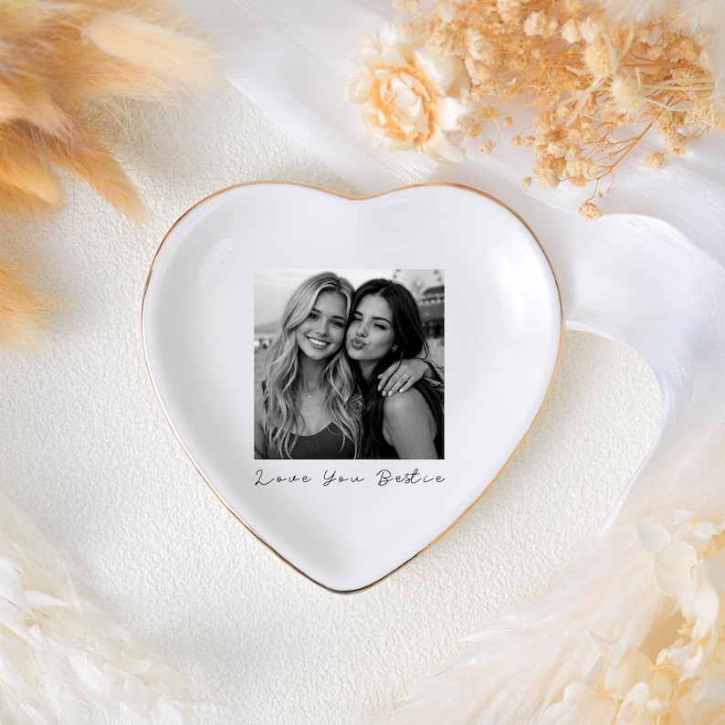 May include: A heart-shaped white ceramic trinket dish with a gold rim. The dish features a black and white photo of two smiling women. The words "Love You Bestie" are written in script below the photo. Decorative elements surround the dish.