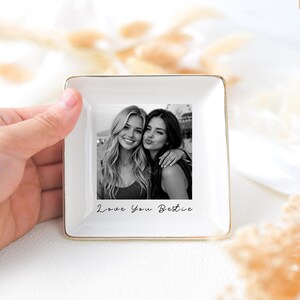 May include: A square, white ceramic trinket dish with a gold rim, featuring a black and white photo of two women. The photo is centered and has the text "Love You Bestie" below it. The dish is held by a hand.
