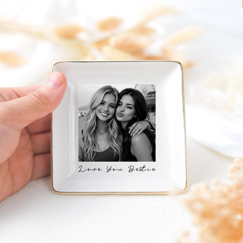 May include: A square, white ceramic trinket dish with a gold rim, featuring a black and white photo of two women. The photo is centered and has the text "Love You Bestie" below it. The dish is held by a hand.