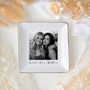 May include: White square ceramic trinket dish with a gold rim. The center features a black and white photo of two women with the text "Love You Bestie". A decorative item for a desk or dresser.
