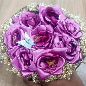 Puede incluir: Un ramo de rosas de satén de color púrpura intenso dispuestas en un grupo circular, acentuado con un pequeño adorno de mariposa plateada. El borde exterior del arreglo está rodeado por un borde decorativo o follaje dorado metálico.
