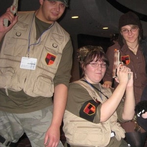 May include: Three people dressed in camouflage vests and holding toy guns. The vests have patches with a red and black design. The people are posing for a photo.
