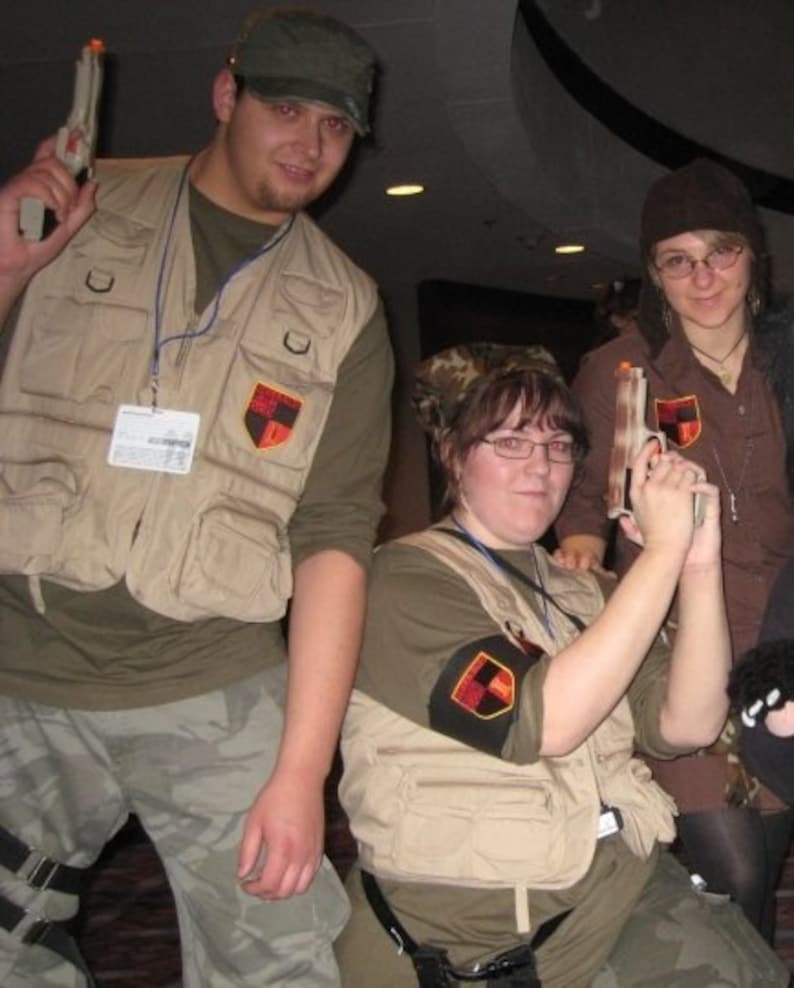 May include: Three people dressed in camouflage vests and holding toy guns. The vests have patches with a red and black design. The people are posing for a photo.