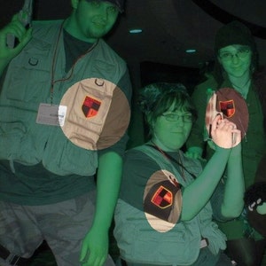 May include: Two people wearing green and brown vests with patches. The vests have a black, red, and yellow shield design. The person on the left is holding a green toy gun.