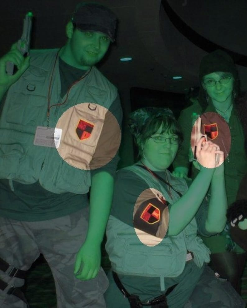 May include: Two people wearing green and brown vests with patches. The vests have a black, red, and yellow shield design. The person on the left is holding a green toy gun.