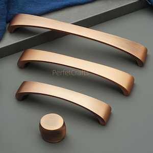 May include: A collection of bronze-toned cabinet handles and a knob, displayed on a grey surface with a blue cloth. The set includes three curved handles of varying lengths and a circular knob. The hardware is for cabinets.