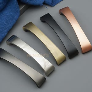 Pode incluir: Um conjunto de seis puxadores de armário em vários acabamentos metálicos: prata, ouro, preto e cobre. Os puxadores têm um design curvo e estão dispostos sobre uma superfície cinzenta.