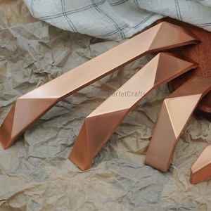 May include: A collection of copper-toned cabinet handles and a knob, featuring geometric, faceted designs. The hardware is arranged on a textured, brown paper surface, with a wooden serving tray and a white and grey checked cloth in the background.