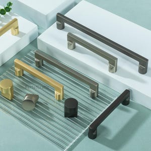 May include: A collection of cabinet hardware in various metallic finishes, including gold, silver, and dark bronze. The assortment features both bar pulls and knob styles, displayed on white platforms and a textured glass surface.