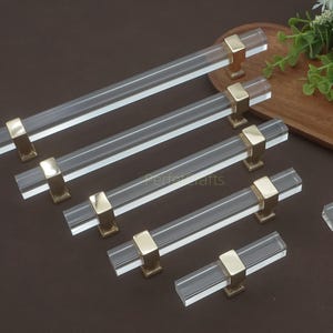 Op de afbeelding: Een verzameling heldere acryl kastgrepen met goudkleurige metalen bases. De grepen zijn rechthoekig en variëren in grootte, gerangschikt op een bruin oppervlak. De hardware is ontworpen voor meubels en woondecoratie.
