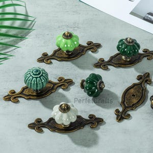 May include: A collection of decorative cabinet knobs in various colors, including green, white, and black. The knobs have a floral design and are mounted on ornate, antique-style bronze backplates. The image also includes a blank paper with the word "Chokla" on it.