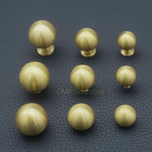 May include: Nine brass cabinet knobs of different sizes are displayed on a dark, textured surface. Each knob is spherical with a small, round base. The brass has a matte finish, reflecting the light.