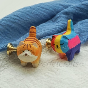 May include: Two wooden cat-shaped cabinet knobs. One is orange with brown stripes, the other is painted in rainbow colors. Both have brass hardware and are set against a blue cloth background. The text "PerfecCrafts" is visible.