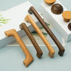 Può includere: Una collezione di ferramenta per mobili in legno, tra cui quattro maniglie e tre pomelli. Le maniglie variano di colore dal marrone chiaro allo scuro. I pomelli sono rotondi e cilindrici, con una finitura a venatura naturale del legno. La ferramenta è esposta su una superficie bianca.