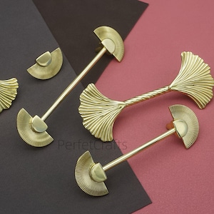 May include: Gold-colored cabinet hardware set featuring ginkgo leaf and fan designs. Includes two long pulls, two small knobs, and two leaf-shaped accents. The hardware is displayed on a dark brown and pink background.