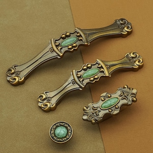 May include: An assortment of antique-style cabinet hardware in bronze and green. The set includes two long handles, a smaller handle, and a round knob. Each piece features an oval, green stone inlay and ornate detailing.