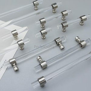 May include: Clear acrylic cabinet pulls with polished nickel hardware. The pulls are cylindrical and have a modern design. The hardware is round and connects the acrylic rod to the cabinet. The image shows several pulls of varying lengths.