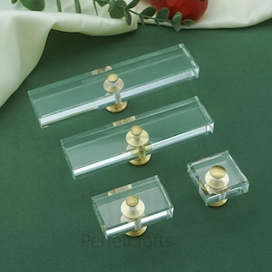 Peut inclure: Boutons de meuble rectangulaires et carrés transparents avec quincaillerie de couleur laiton. Les boutons sont disposés sur une surface vert foncé, avec une figurine d'oiseau rouge et de la verdure en arrière-plan. Les boutons sont de tailles variables.