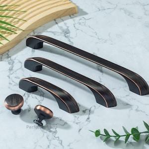 Op de afbeelding: Een verzameling bronzen meubelbeslag, waaronder drie handgrepen en twee knoppen. De handgrepen hebben een gebogen ontwerp met een donkere afwerking en koperen accenten. De knoppen zijn rond met een vergelijkbare afwerking. Het beslag ligt op een marmeren oppervlak.