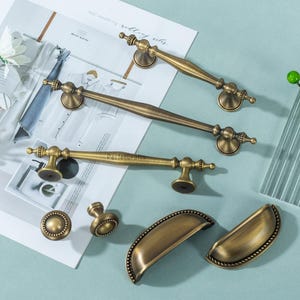 May include: An assortment of antique brass cabinet hardware, including handles, knobs, and pulls. The handles vary in length, with decorative detailing on the ends. The hardware is arranged on a light blue surface, with a magazine and floral accents in the background.