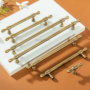May include: Several antique brass cabinet pulls and handles are arranged on a white surface. The hardware features a T-bar design with a vintage aesthetic. The pulls vary in length, showcasing a range of sizes for different cabinet applications.