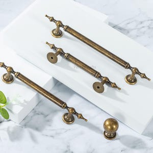 May include: Antique bronze cabinet hardware set. The set includes three long handles and one round knob. The handles have a decorative design with a central bar and ornate ends. The knob has a matching finish and a spherical shape. The hardware is displayed on a white surface.