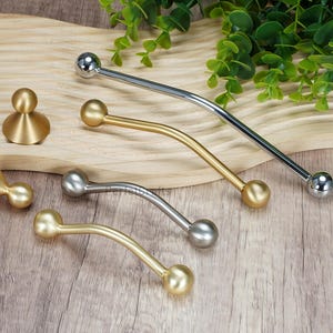 Puede incluir: Una variedad de tiradores y pomos para armarios en acabados dorado, plateado y gris. Los tiradores tienen un diseño curvo con extremos en forma de bola. Los pomos son dorados y tienen una forma distintiva. Medidas en centímetros.