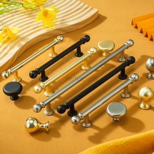 May include: A collection of cabinet hardware in various styles and finishes. Includes gold, black, and silver handles and knobs. The hardware is displayed on a mustard-colored surface, with some yellow flowers in the background.