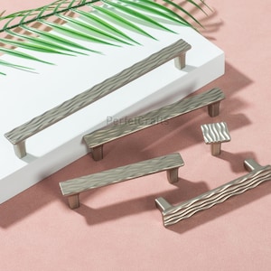 May include: A selection of silver cabinet handles and a knob with a textured, wave-like pattern. The hardware is displayed on a white surface against a pink background, with a green leaf in the upper left corner. The text "PerfectCrafts" is visible.