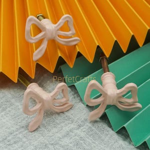 May include: Three light pink bow-shaped cabinet knobs with brass hardware. The knobs are arranged on a textured surface with a backdrop of folded yellow and green paper. The text "PerfetcCrafts" is visible.