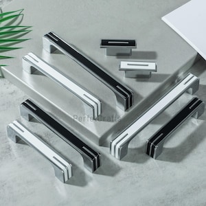 May include: A collection of modern cabinet hardware, including silver-toned handles and knobs. The handles feature a sleek design with black or white center accents and silver stripes. The knobs have matching black or white center accents.
