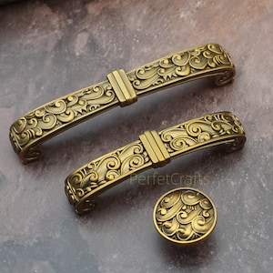 Peut inclure: Ensemble de quincaillerie de meuble en laiton antique. L'ensemble comprend deux poignées incurvées et un bouton rond, tous avec un design orné et tourbillonnant. La quincaillerie est en métal et a une esthétique vintage.