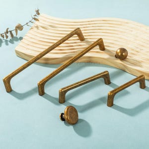 Puede incluir: Un conjunto de herrajes de latón antiguo para armarios, que incluye tiradores y pomos. Los herrajes tienen un acabado cálido y envejecido y están dispuestos sobre una superficie azul claro con un elemento de madera. El texto "PerfecCrafts" es visible.