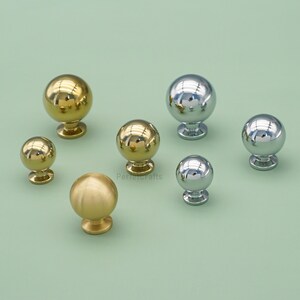 May include: A selection of spherical cabinet knobs in gold and silver finishes. The knobs vary in size and have a polished, reflective surface. The knobs are displayed on a pale green background.