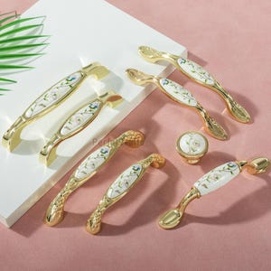 Peut inclure: Ensemble de poignées de meubles dorées et un bouton, avec des incrustations en céramique blanche ornées de motifs floraux bleus, verts et violets. Les poignées ont une forme incurvée et le bouton un motif floral assorti.