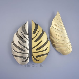 May include: Three decorative leaf-shaped cabinet pulls in metallic finishes. Two pulls are paired, one silver and one gold, with a matching gold pull to the right. The pulls have a textured, ridged design against a blue background.