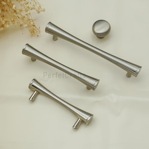 May include: A collection of brushed nickel cabinet hardware, featuring three elongated handles and a round knob. The handles have a tapered design, and the knob is a simple, rounded shape. The hardware is displayed on a light-coloured surface.