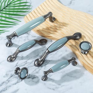 以下が含まれることがあります： キャビネットハードウェアのコレクション。ガンメタル仕上げのハンドルと、ライトブルーのセラミックインサート。ノブは、お揃いのライトブルーのセラミックセンター。木製の表面に表示されています。