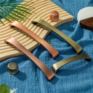 Może przedstawiać: Kolekcja okuć do szafek w różnych metalicznych wykończeniach, w tym miedzi, mosiądzu i brązu. Na zdjęciu widoczne są uchwyty i gałki. Widoczny jest tekst "PerfectCrafts". Wymiary w centymetrach.