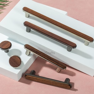 May include: A collection of wooden cabinet hardware, including pulls and knobs. The pulls have a rectangular wooden center with silver-toned metal ends. The knobs are round with a wooden center and silver-toned metal base. The hardware is displayed on a white surface.